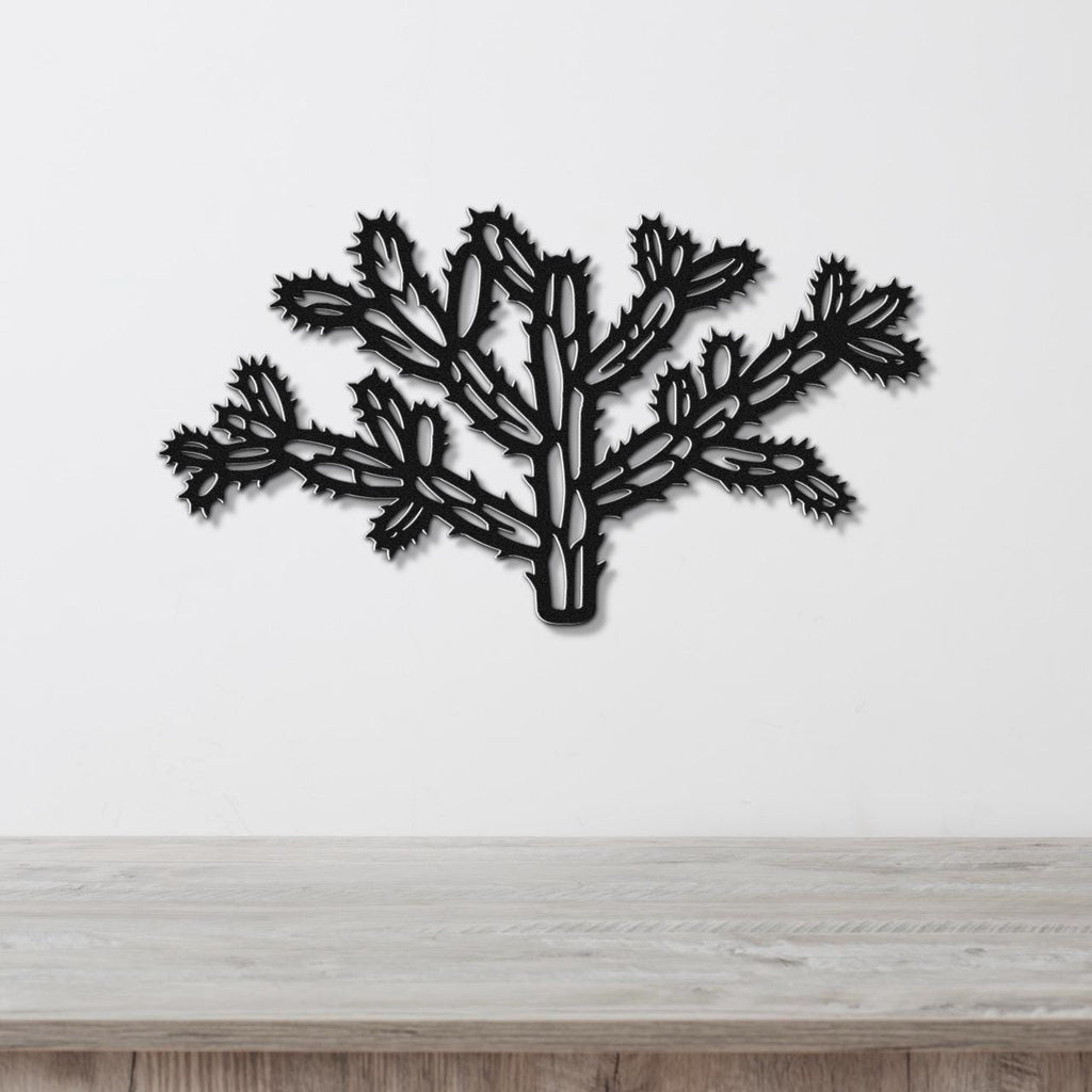 Black metal wall art cutout of a cholla cactus with intricate branching and spiny segments, mounted on a white wall above a light wood surface.