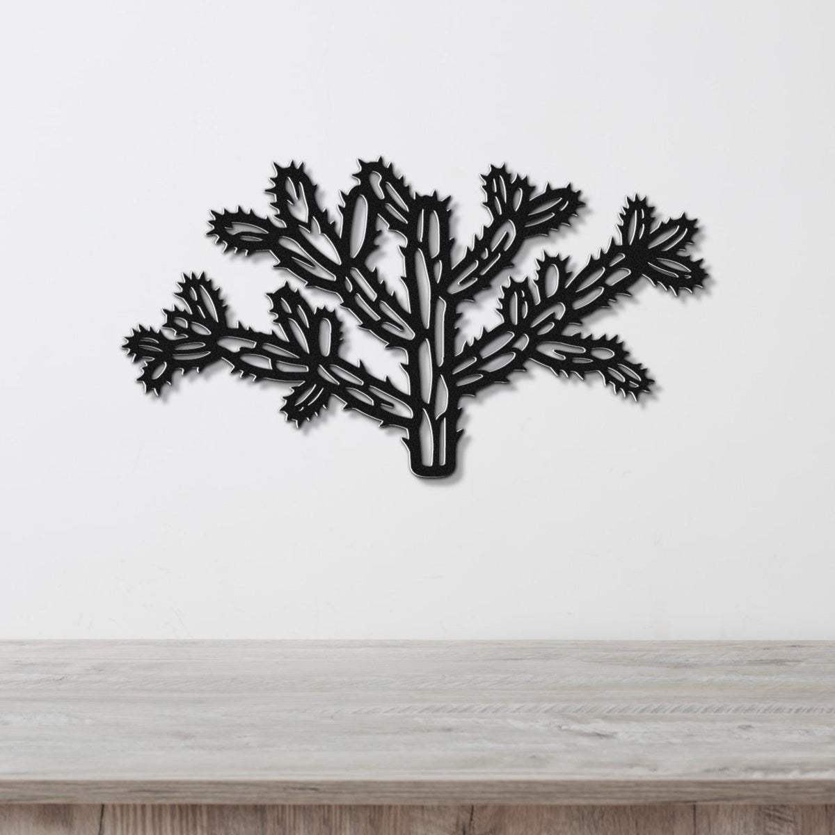 Black metal wall art cutout of a cholla cactus with intricate branching and spiny segments, mounted on a white wall above a light wood surface.