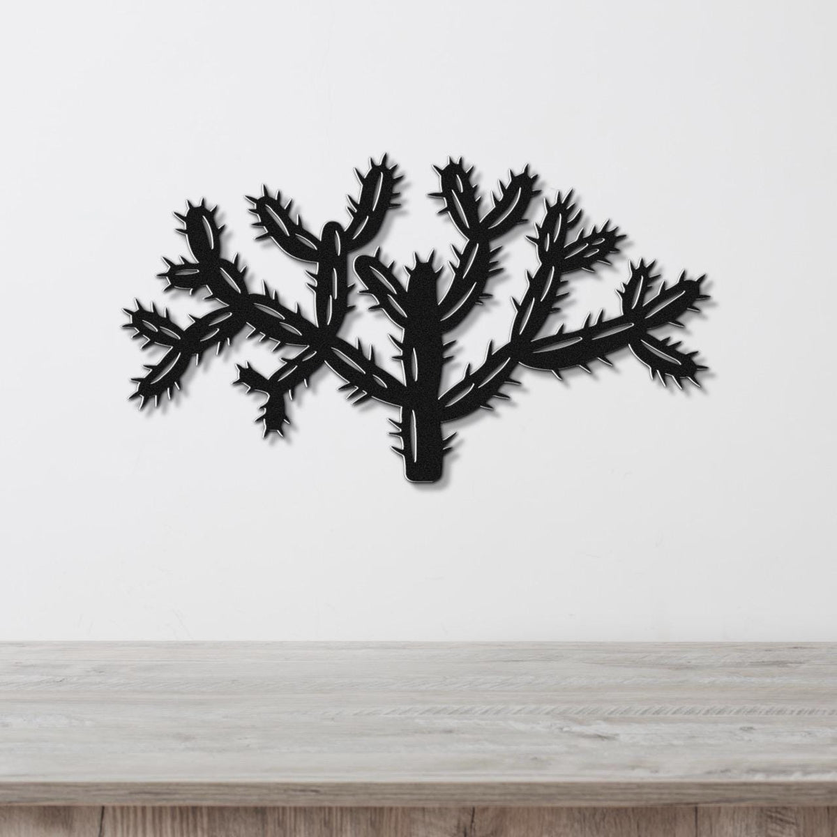 Black metal wall art cutout of a cholla cactus with textured spine details, mounted on a white wall above a light wood surface.