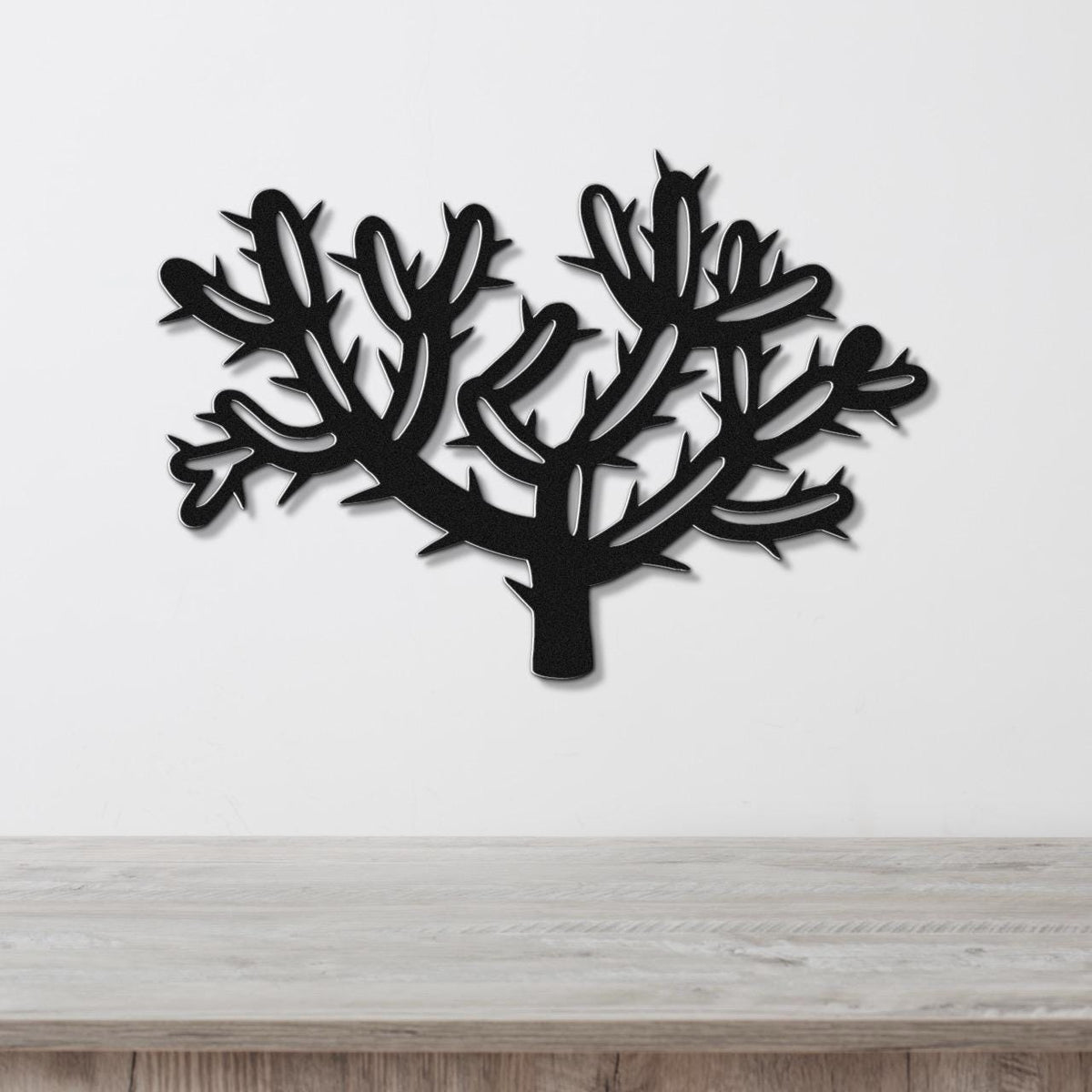 Black metal wall art cutout of a cholla cactus with branching arms and spines, mounted on a white wall above a light wood surface.