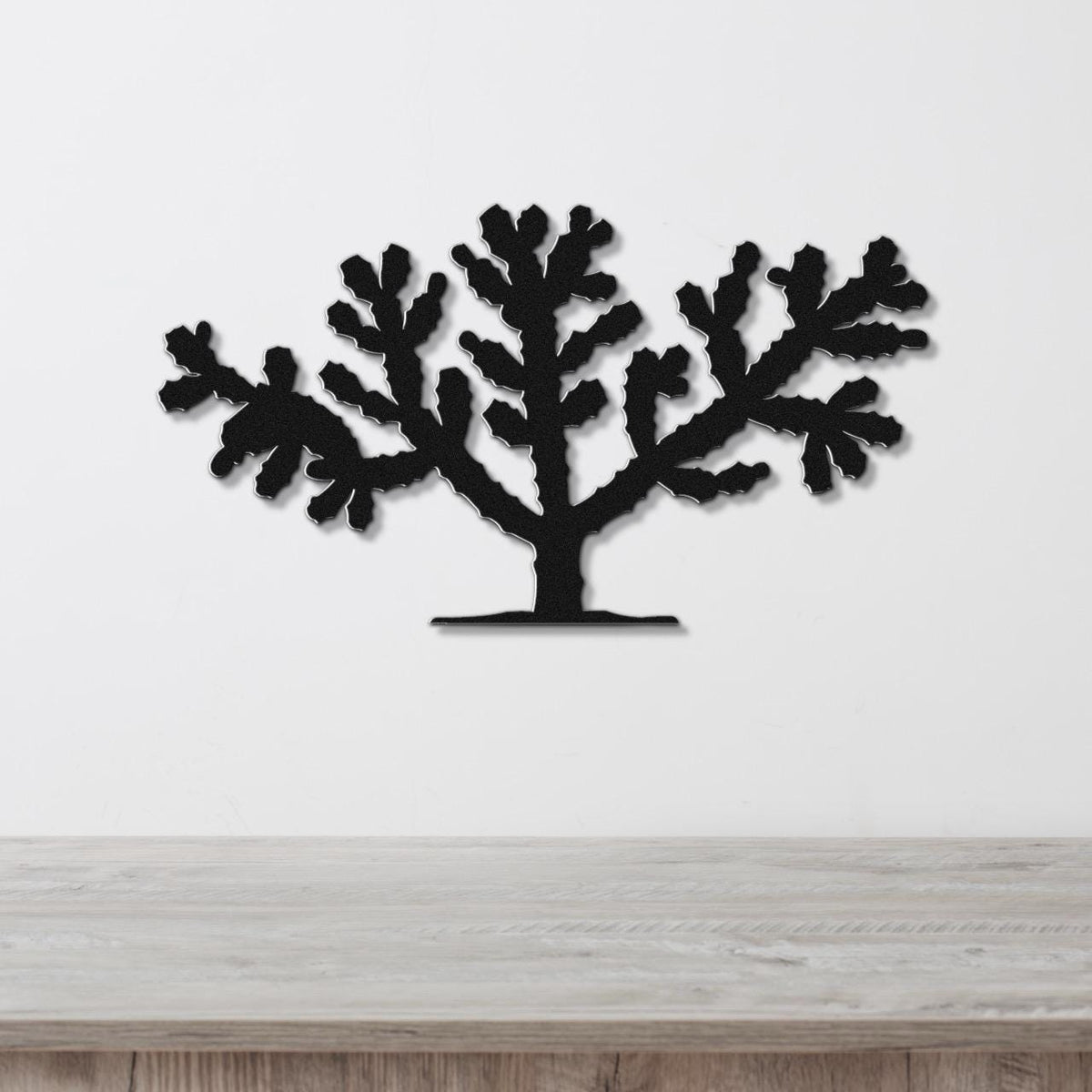 Black metal wall art silhouette of a cholla cactus with branching arms, mounted on a white wall above a light wood surface.