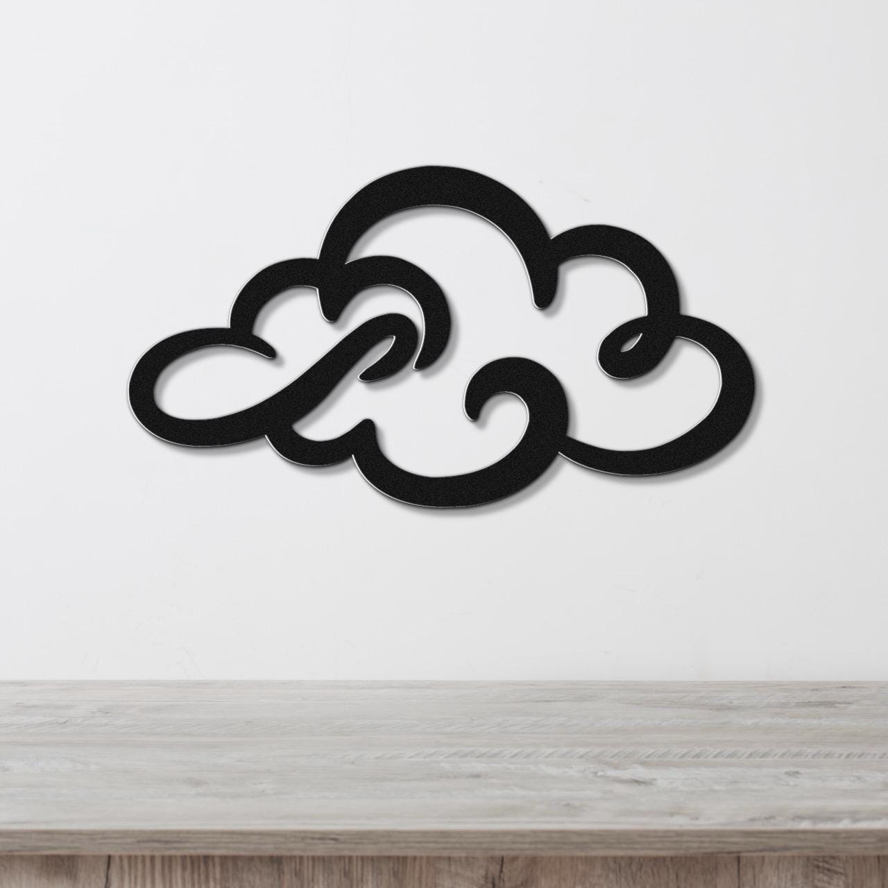 Black metal cloud wall art with swirling cutout design, mounted on a white wall above a light wood surface.