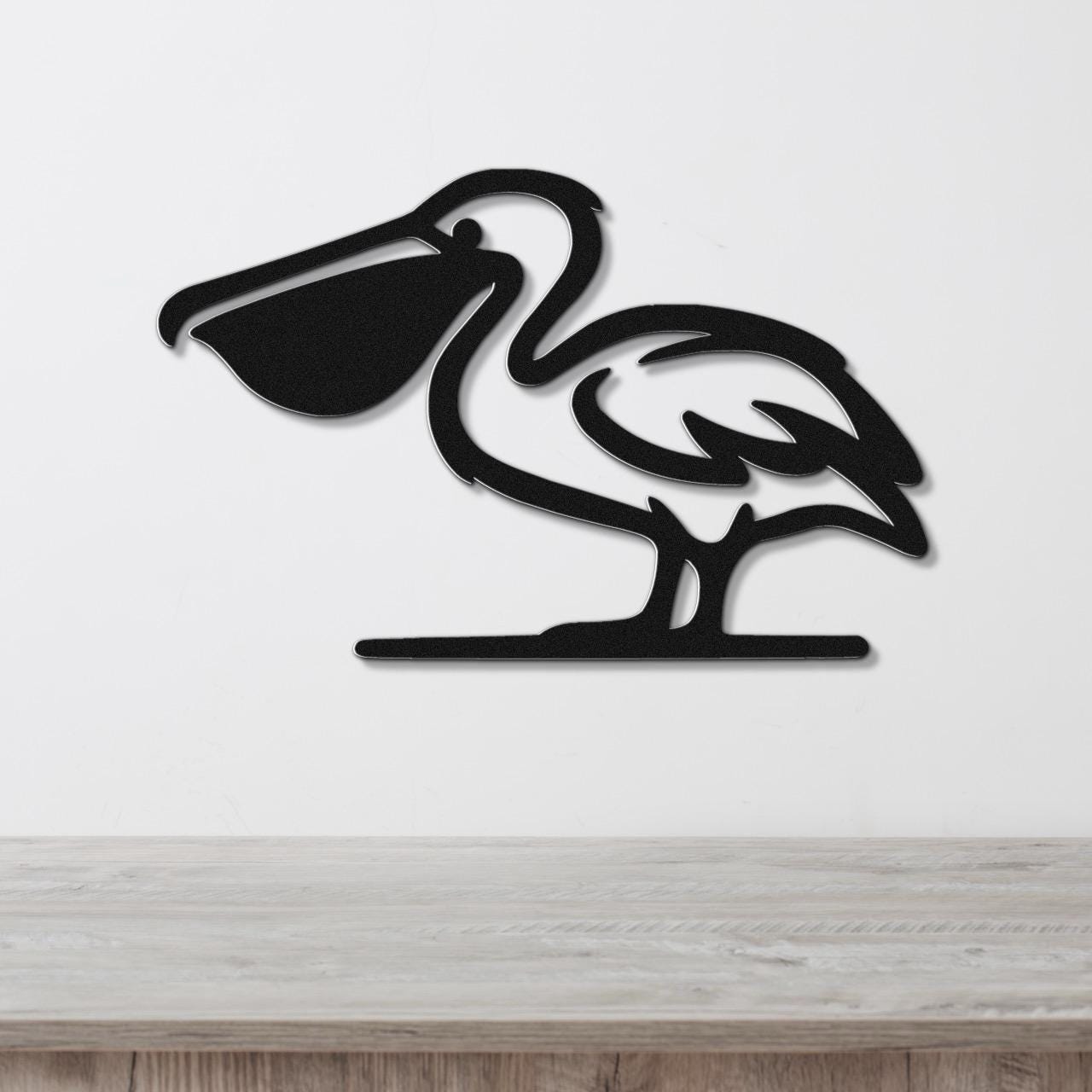 Black metal pelican wall art with a cutout silhouette design, mounted on a white wall above a light wood surface.