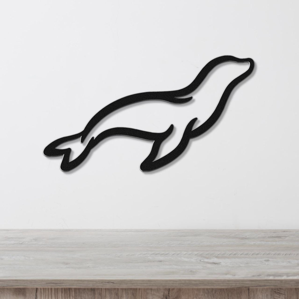 Black metal sea lion wall art with a sleek silhouette design, mounted on a white wall above a light wood surface.