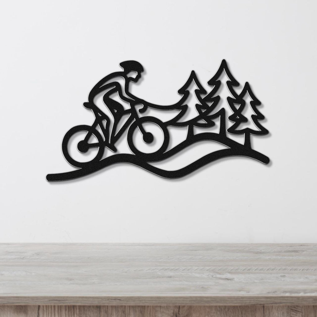 Black metal wall art silhouette of a cyclist on a trail with pine trees, mounted on a white wall above a wooden surface.
