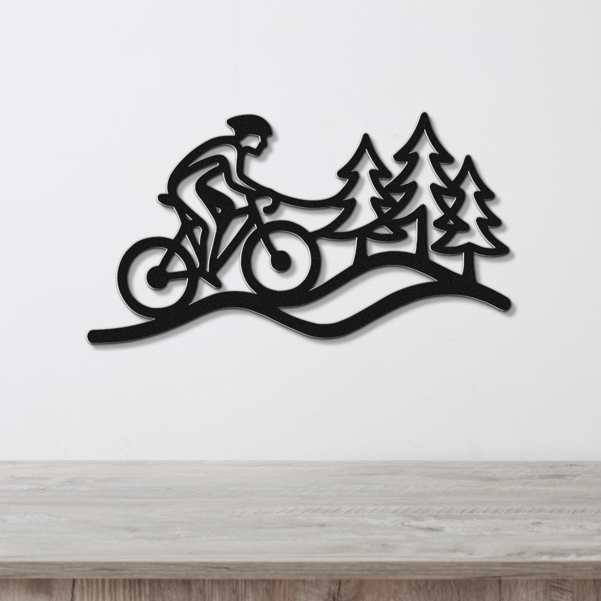 Black metal wall art silhouette of a cyclist on a trail with pine trees, mounted on a white wall above a wooden surface.