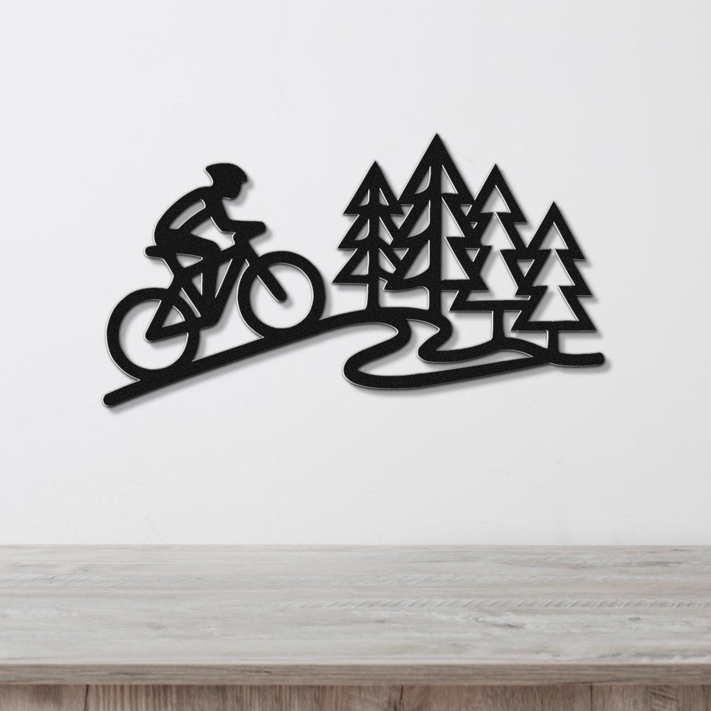 Black metal wall art featuring a cyclist silhouette on a winding path beside pine trees, mounted on a white wall above a wooden surface.
