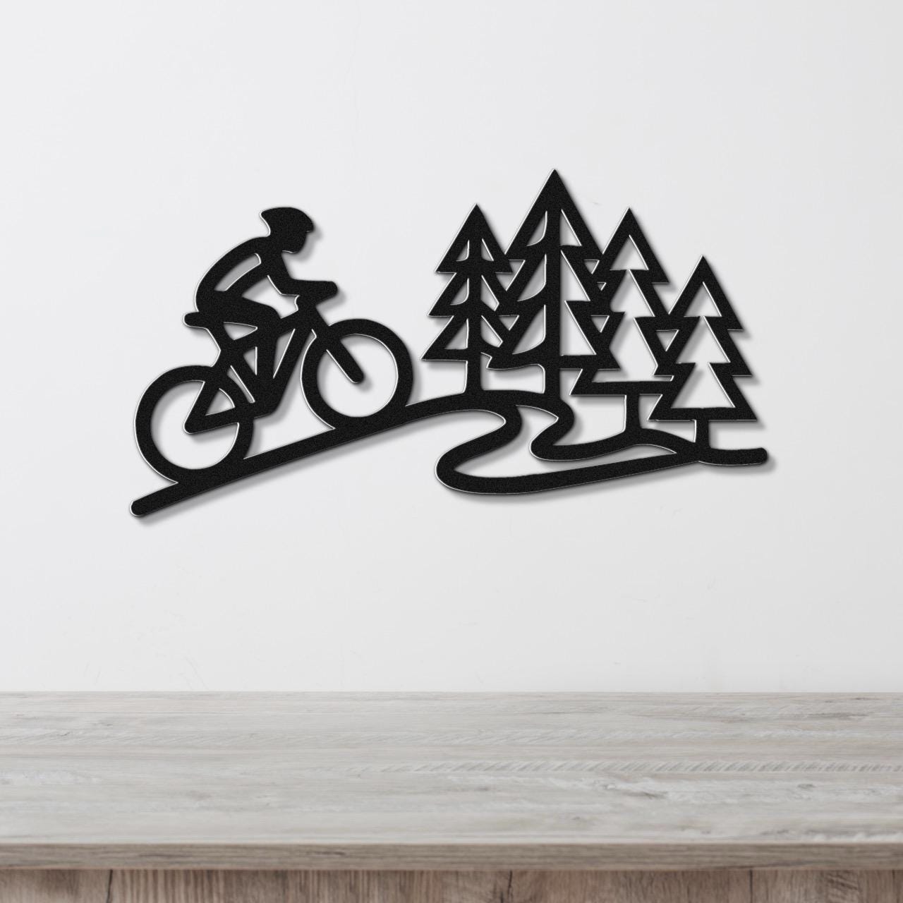 Black metal wall art featuring a cyclist silhouette on a winding path beside pine trees, mounted on a white wall above a wooden surface.