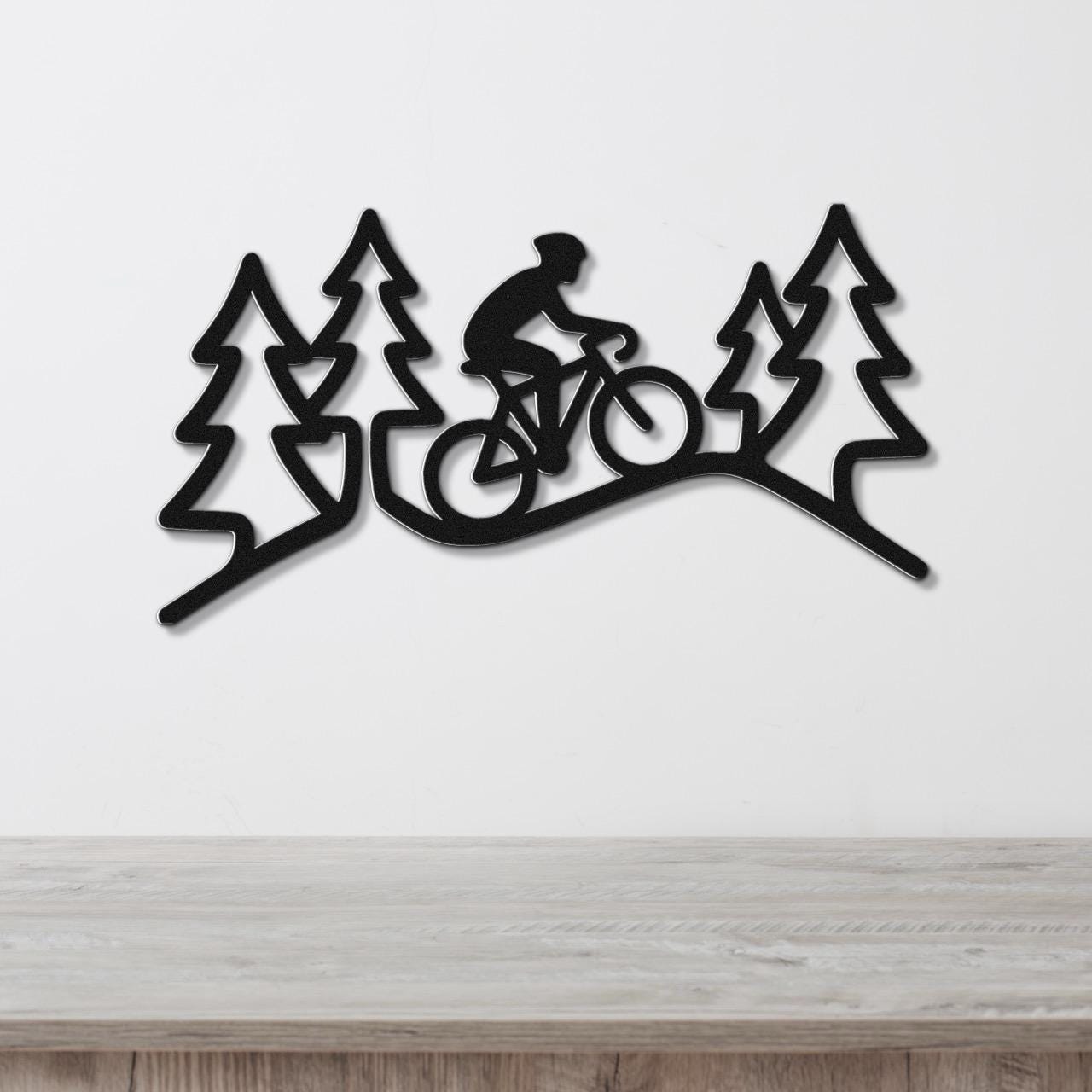Black metal wall art featuring a cyclist silhouette riding through a pine forest, mounted on a white wall above a wooden surface.