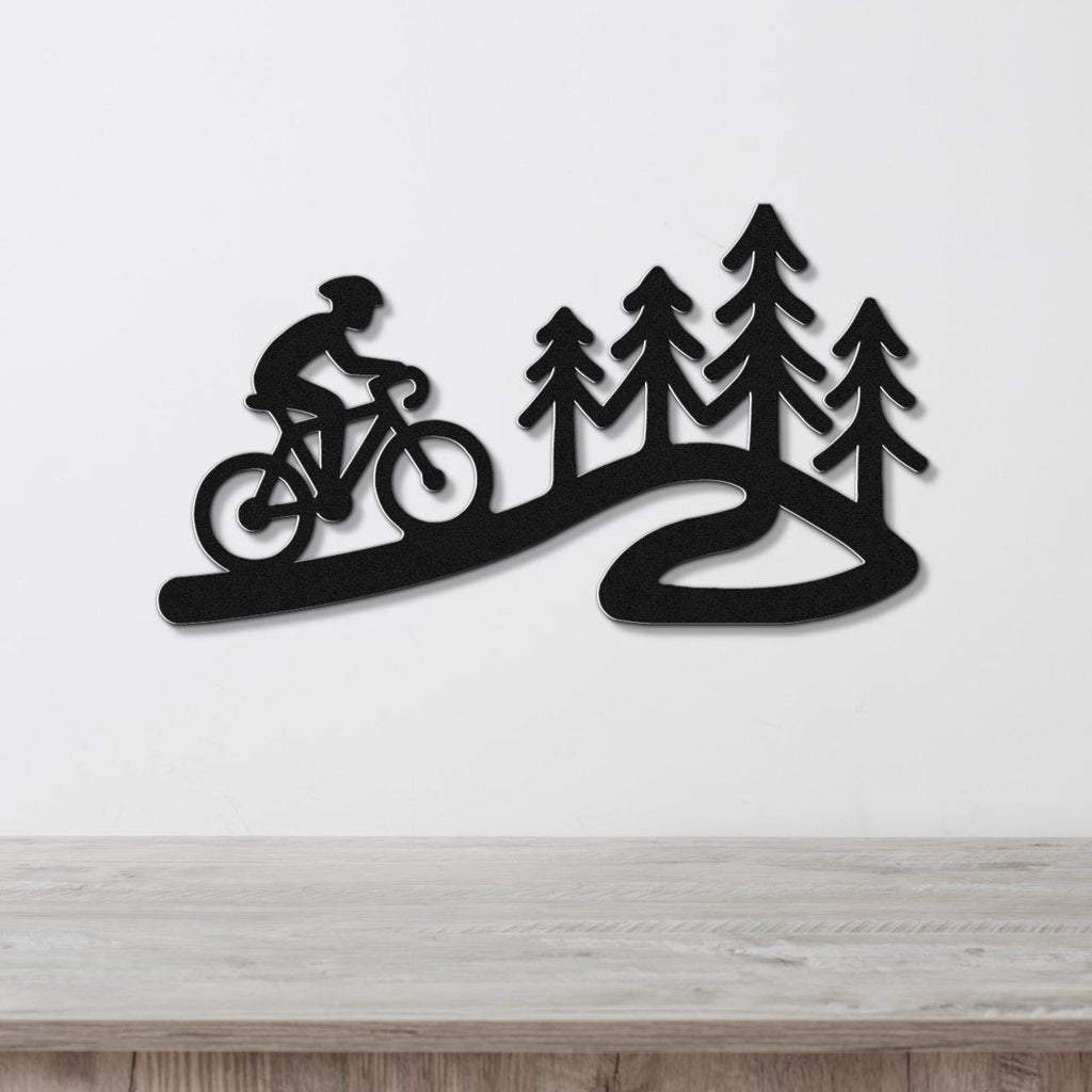 Black metal wall art silhouette of a cyclist on a winding path with pine trees, mounted on a white wall above a wooden surface.