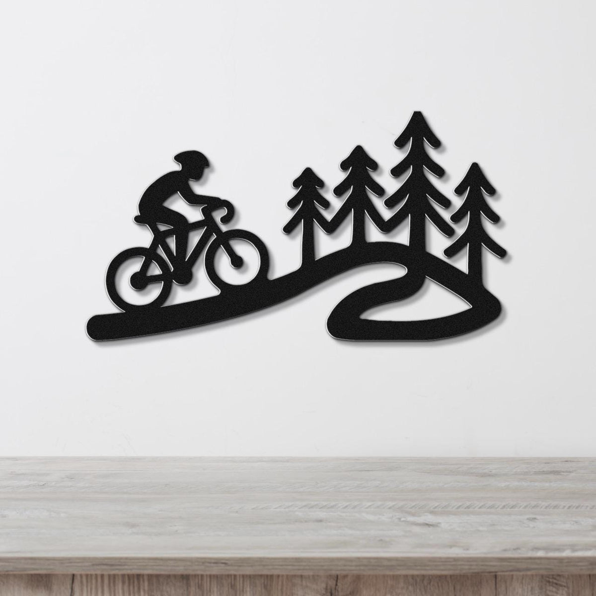 Black metal wall art silhouette of a cyclist on a winding path with pine trees, mounted on a white wall above a wooden surface.