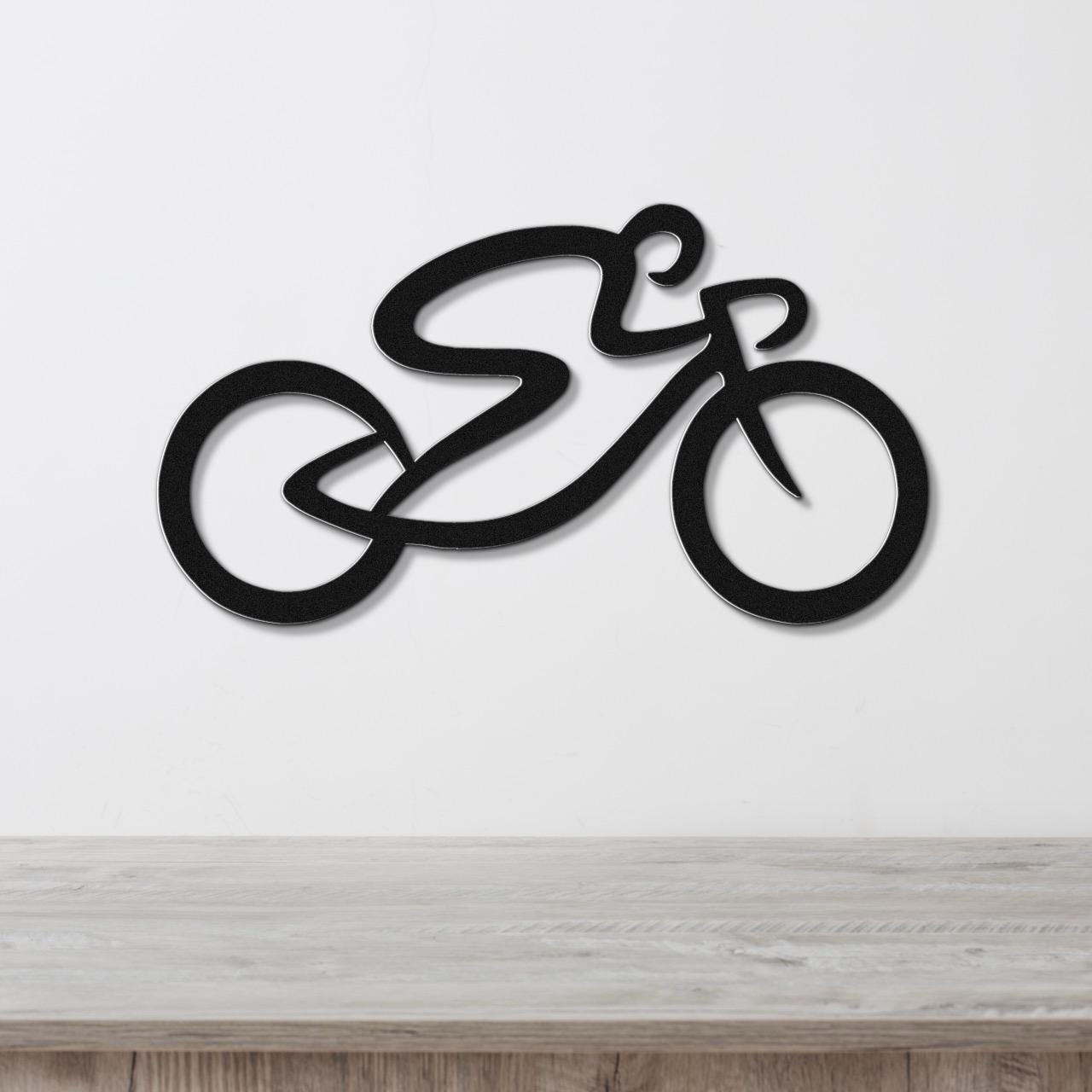 Black metal wall art in the shape of a stylized bicycle with flowing lines, mounted on a white wall above a light wood surface.