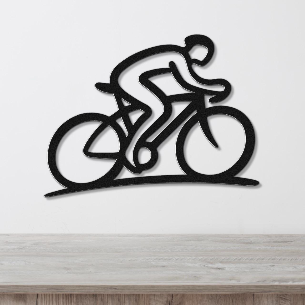Black metal wall art featuring a dynamic cyclist silhouette with internal cutouts, mounted on a white wall above a wooden surface.