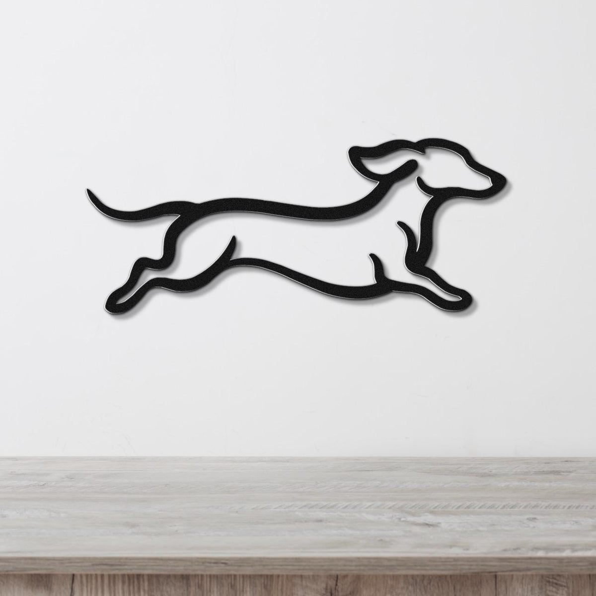 Black metal wall art of a dachshund in a running silhouette, mounted on a white wall above a light wood surface.