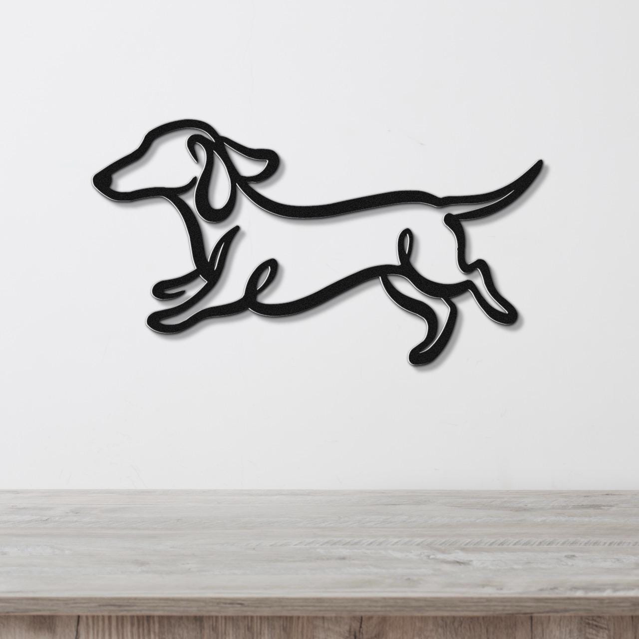 Black metal wall art of a dachshund in a running silhouette, mounted on a white wall above a light wood surface.