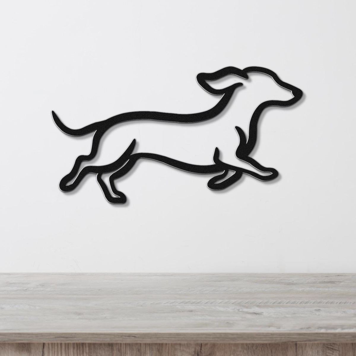 Black metal dachshund wall art in a running silhouette, mounted on a white wall above a light wood surface.