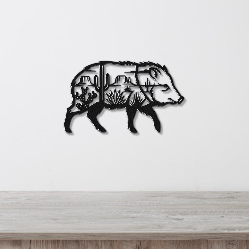 Black metal wall art in the silhouette of a javelina, with internal cutouts of cacti and desert landscape, mounted on a white wall above a wooden surface.