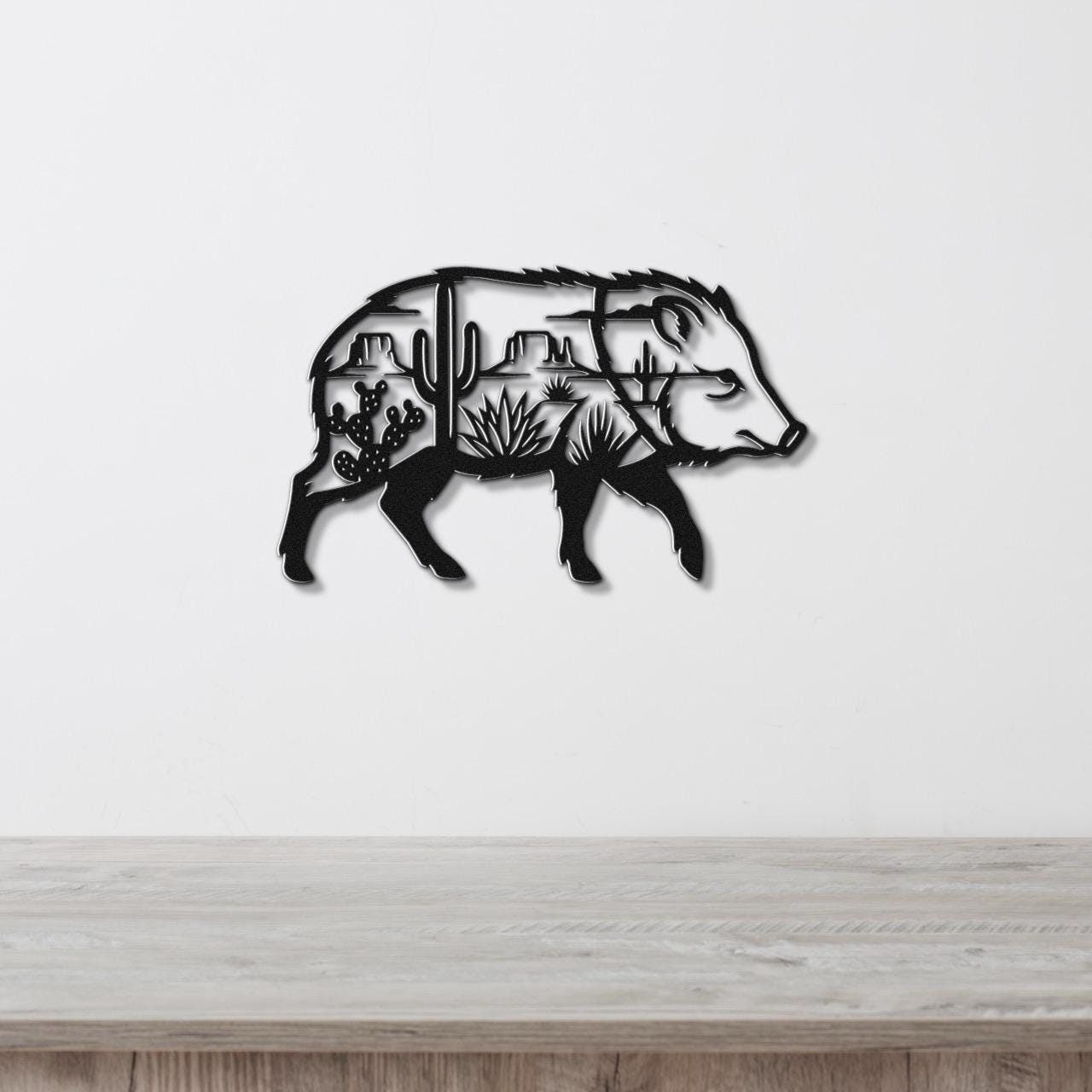 Black metal wall art in the silhouette of a javelina, with internal cutouts of cacti and desert landscape, mounted on a white wall above a wooden surface.