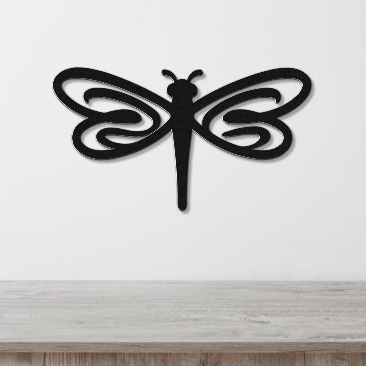 Black metal dragonfly wall art with flowing wing cutouts, mounted on a white wall above a light wood surface.