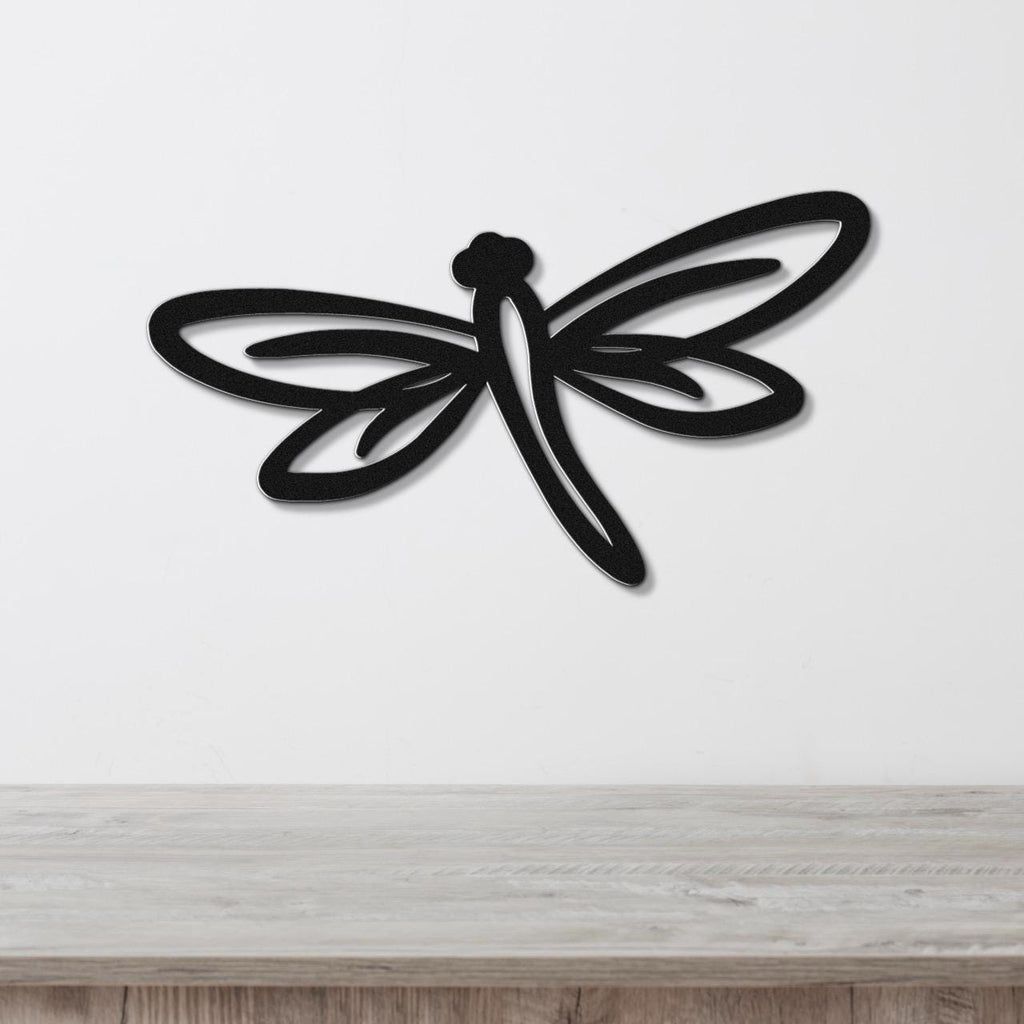 Black metal dragonfly wall art with cutout wings and body, mounted on a white wall above a light wood surface.
