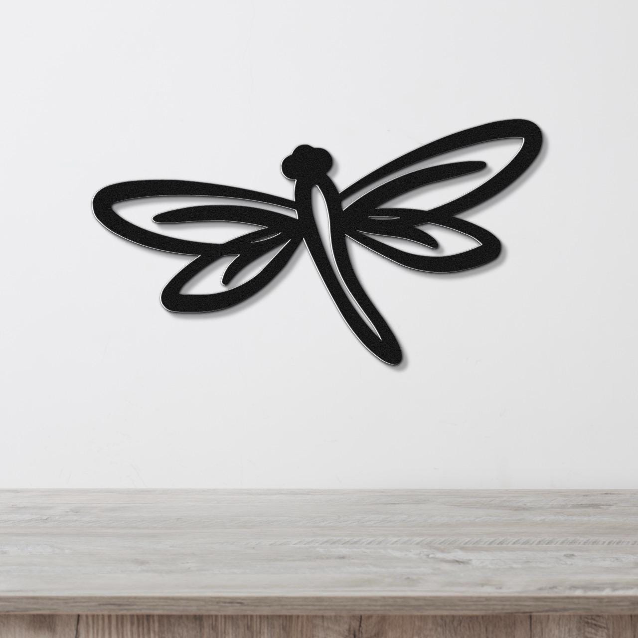Black metal dragonfly wall art with cutout wings and body, mounted on a white wall above a light wood surface.