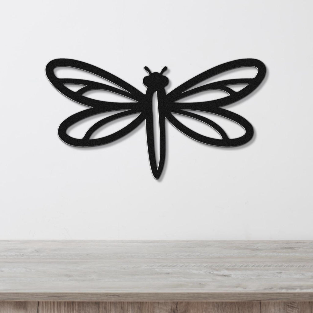 Black metal dragonfly wall art with intricate wing cutouts, mounted on a white wall above a light wood surface.