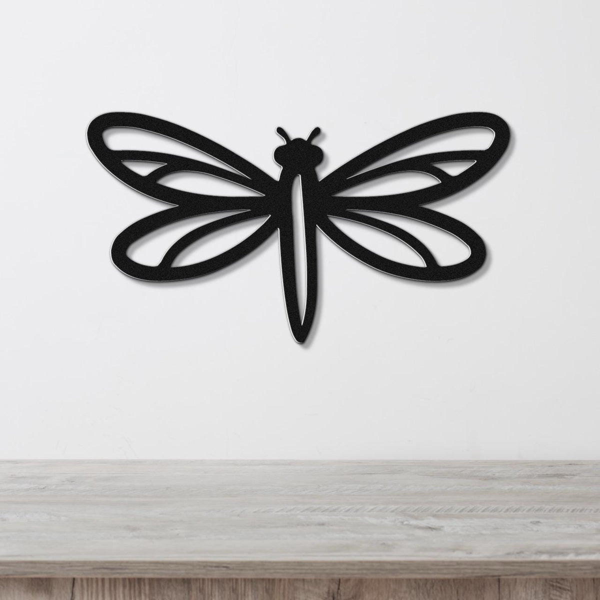 Black metal dragonfly wall art with intricate wing cutouts, mounted on a white wall above a light wood surface.