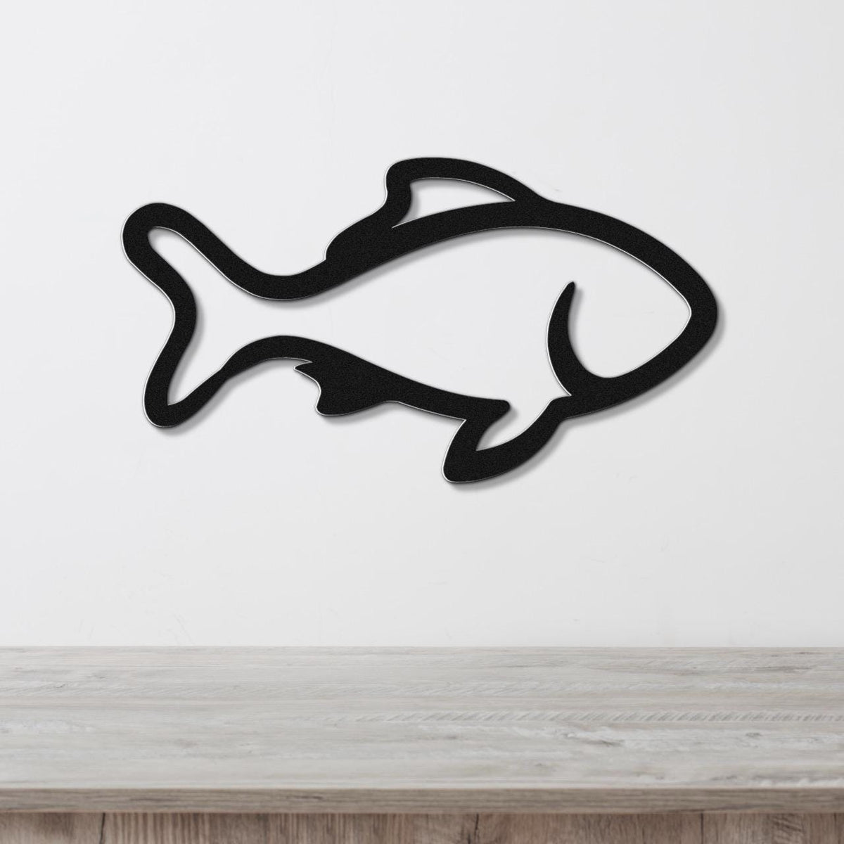 Black metal fish wall art with bold outline and internal cutouts, mounted on a white wall above a light wood surface.
