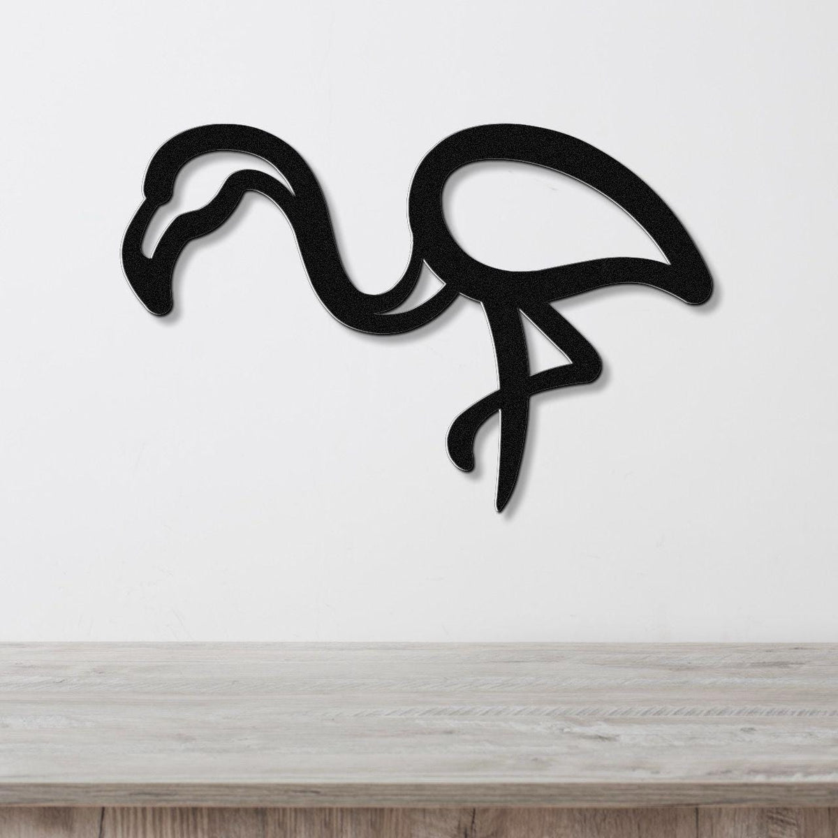 Black metal flamingo wall art with a bold silhouette design, mounted on a white wall above a light wood surface.