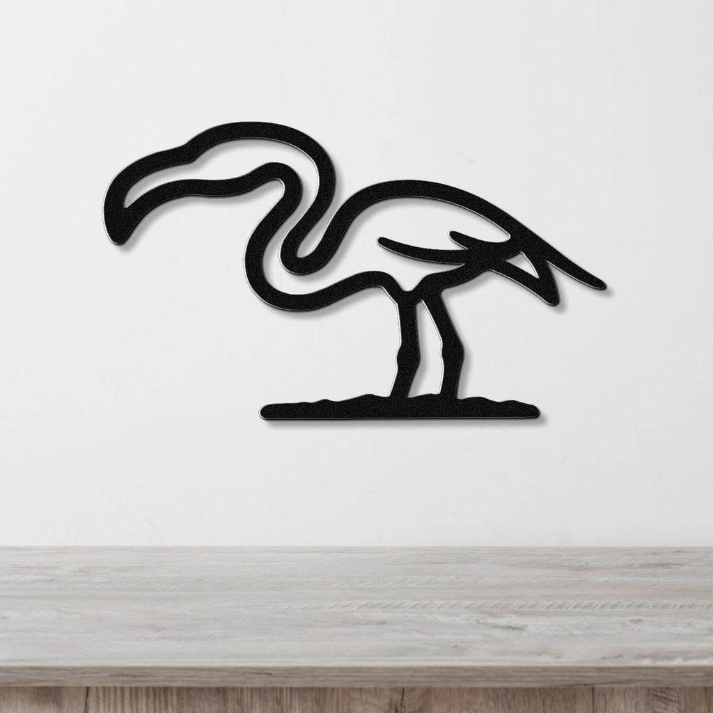 Black metal flamingo wall art with a minimalist silhouette design, mounted on a white wall above a light wood surface.