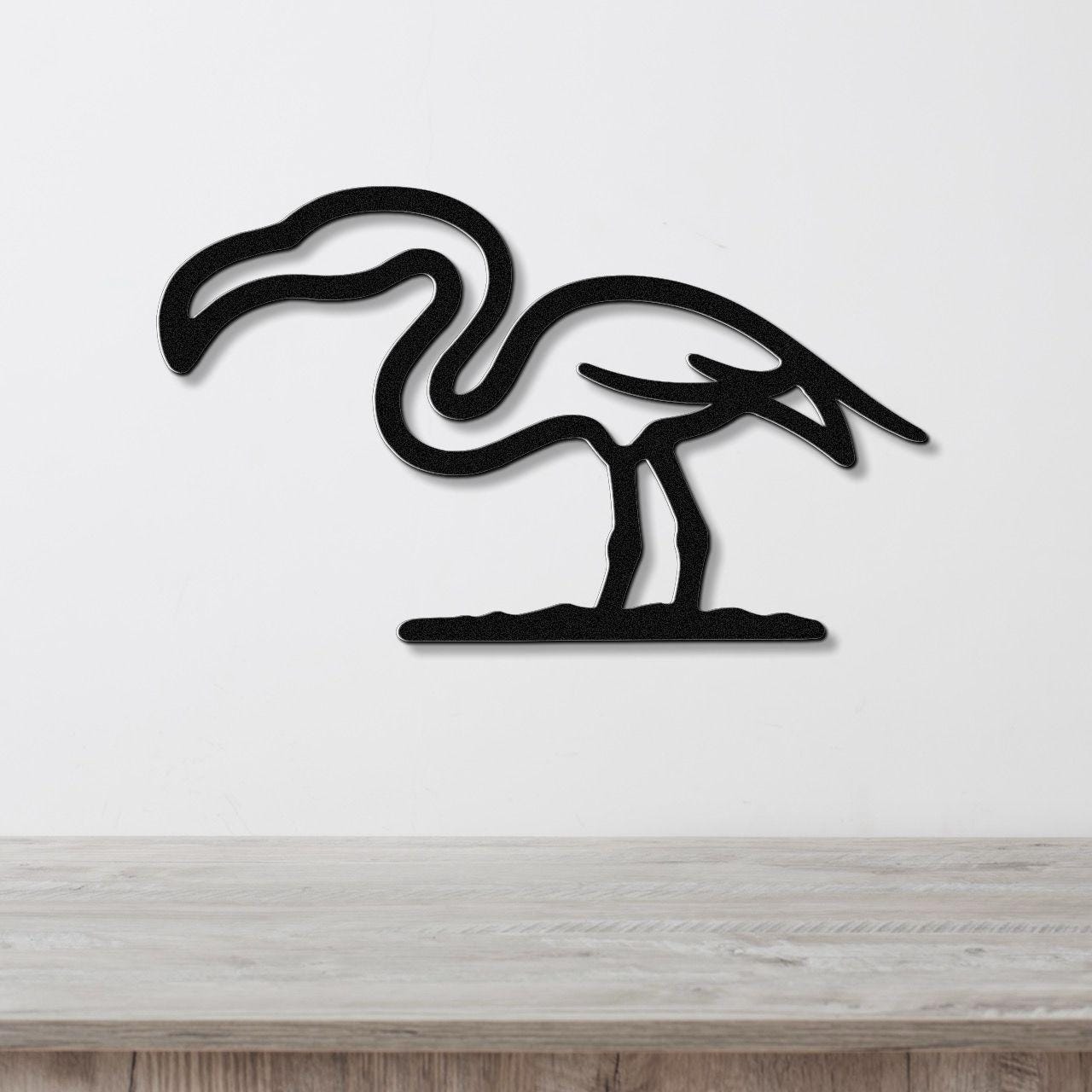 Black metal flamingo wall art with a minimalist silhouette design, mounted on a white wall above a light wood surface.