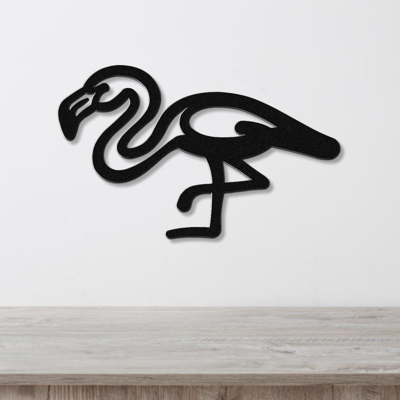Black metal flamingo wall art with a minimalist silhouette design, mounted on a white wall above a light wood surface.