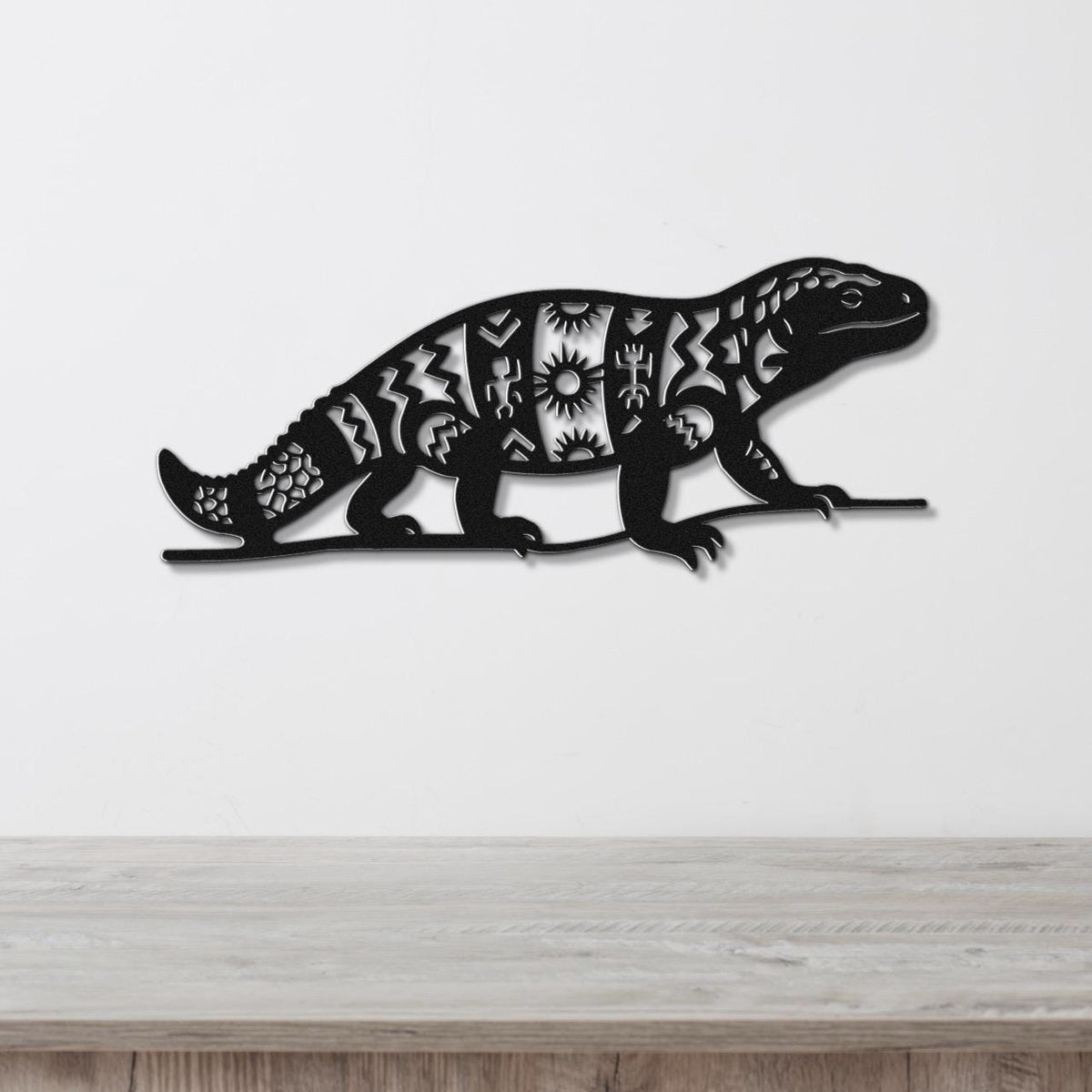 Black metal wall art of a Gila monster with intricate geometric and sunburst cutouts, mounted on a white wall above a wooden surface.