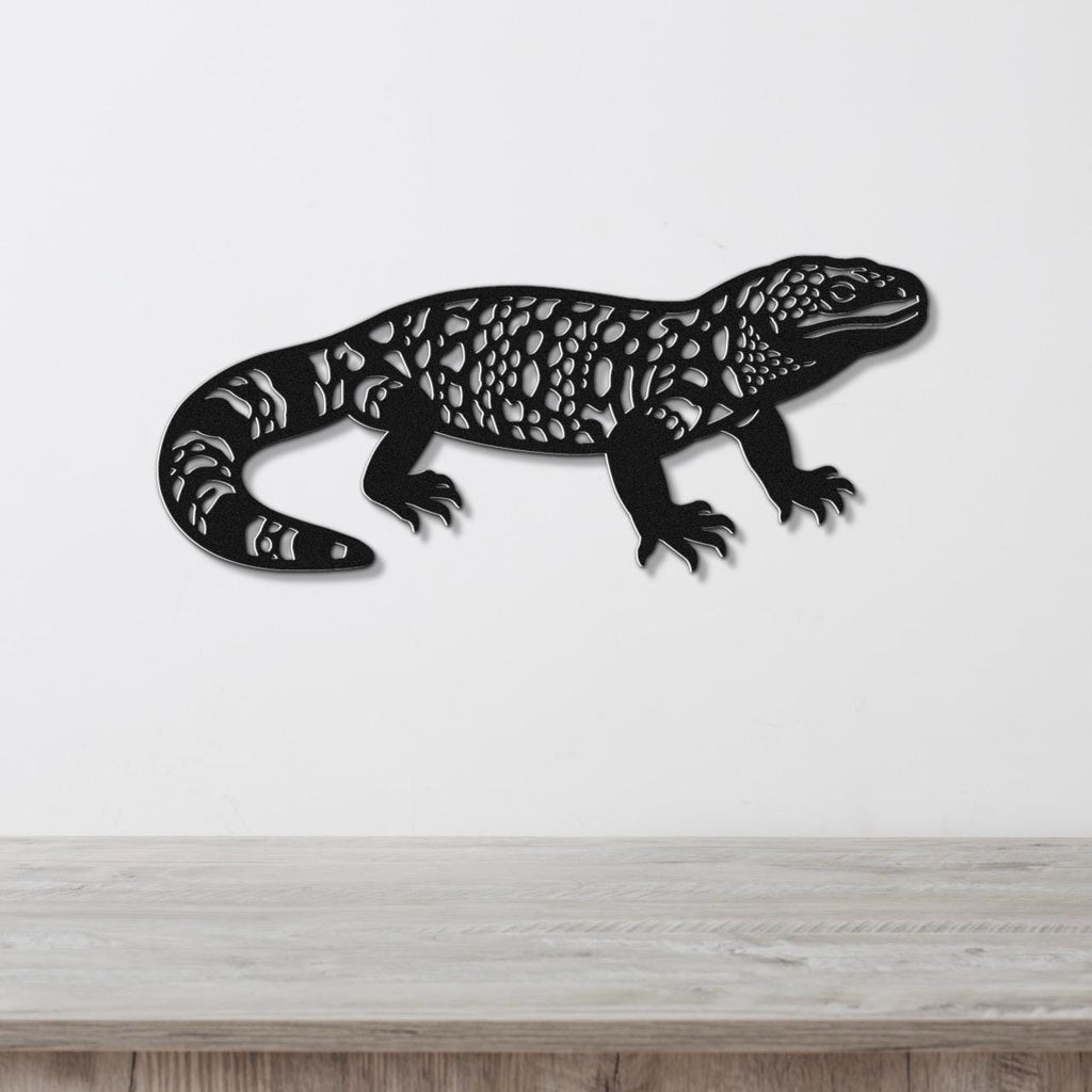 Black metal wall art of a Gila monster with intricate cutout patterns, mounted on a white wall above a wooden surface.