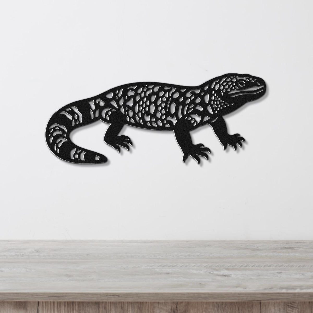Black metal wall art of a Gila monster with intricate cutout patterns, mounted on a white wall above a wooden surface.