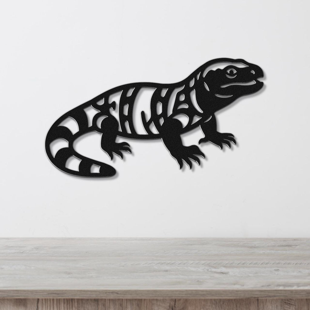 Black metal wall art of a Gila monster with intricate internal cutouts, mounted on a white wall above a light wood surface.