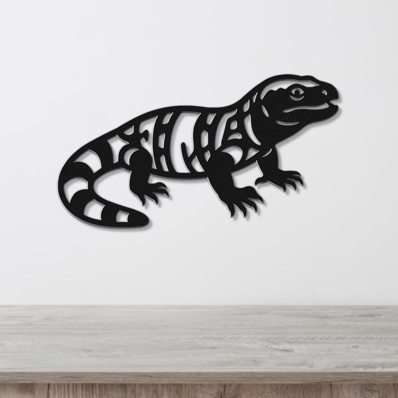 Black metal wall art of a Gila monster with intricate internal cutouts, mounted on a white wall above a light wood surface.