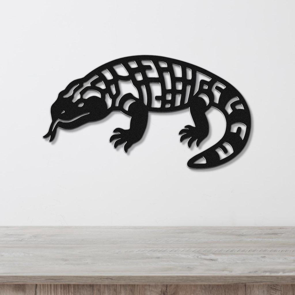 Black metal wall art of a Gila monster with intricate cutout patterns on its body, mounted on a white wall above a wooden surface.