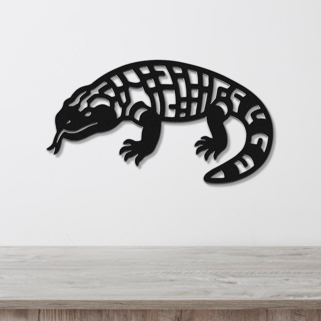 Black metal wall art of a Gila monster with intricate cutout patterns on its body, mounted on a white wall above a wooden surface.