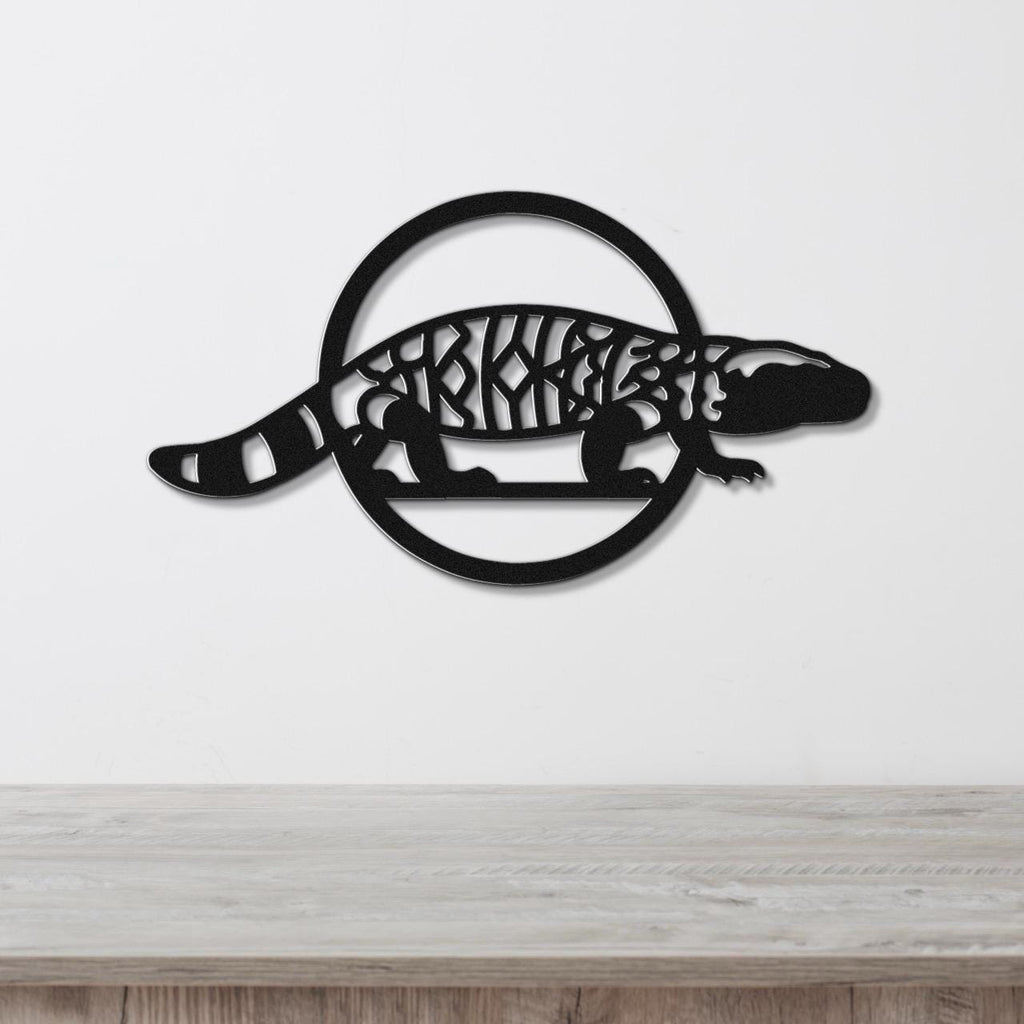 Black metal wall art featuring a Gila monster silhouette with intricate internal cutouts, mounted on a white wall above a light wood surface.