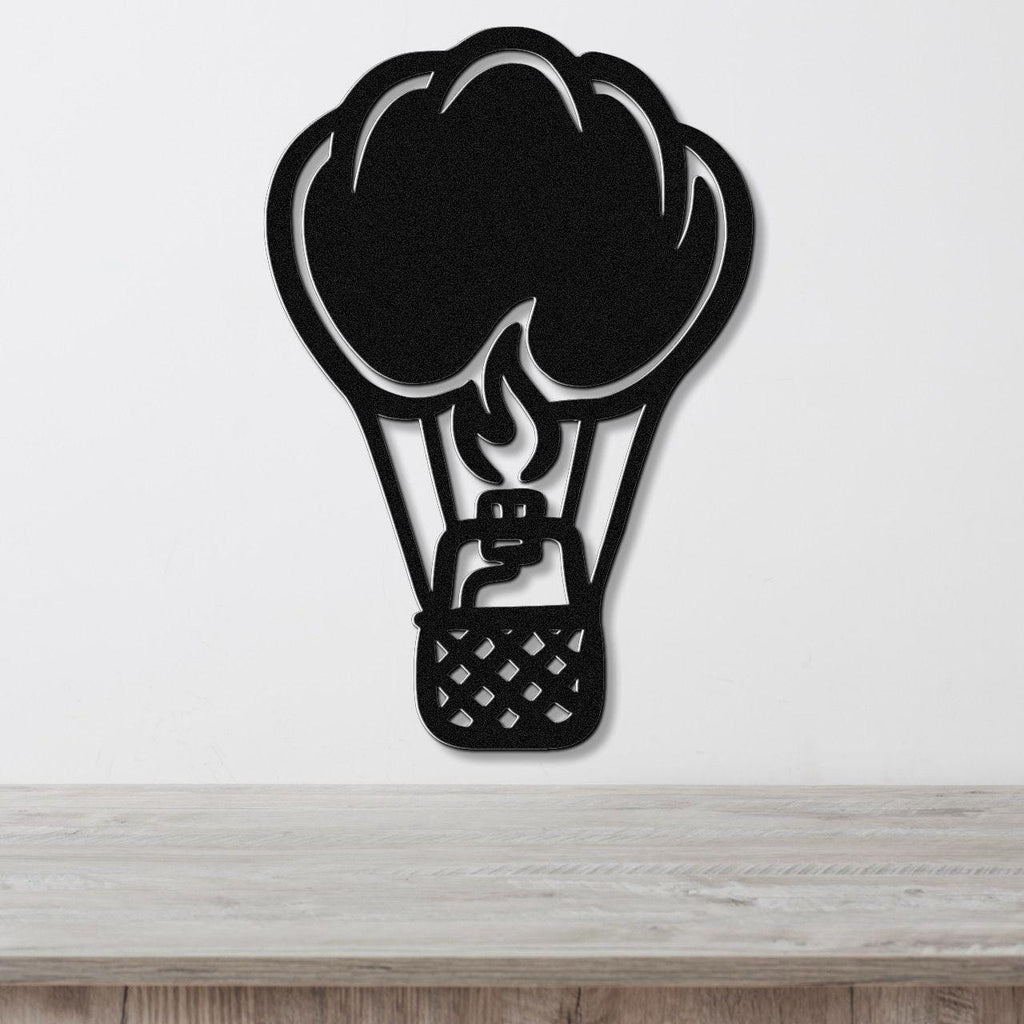 Black metal wall art in the silhouette of a hot air balloon with flame and basket cutouts, mounted on a white wall above a wooden surface.