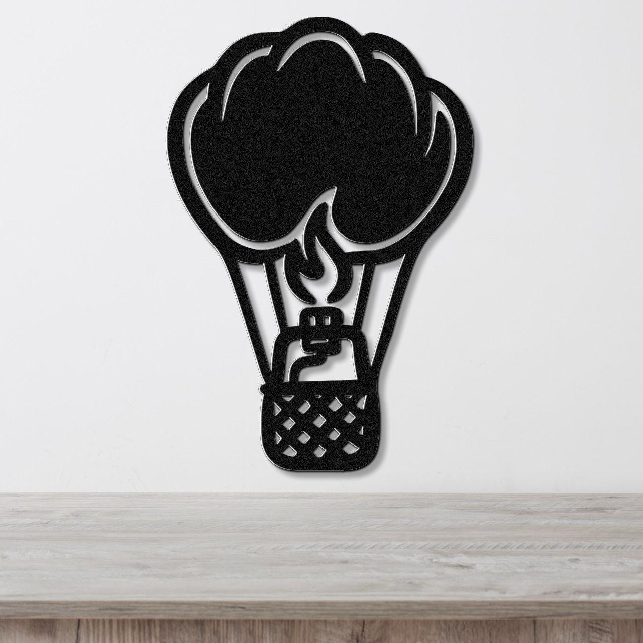 Black metal wall art in the silhouette of a hot air balloon with flame and basket cutouts, mounted on a white wall above a wooden surface.