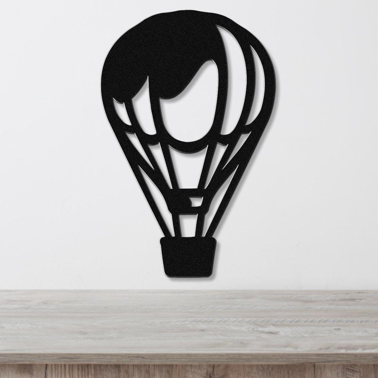 Black metal wall art in the silhouette of a hot air balloon with internal cutout details, mounted on a white wall above a light wood surface.