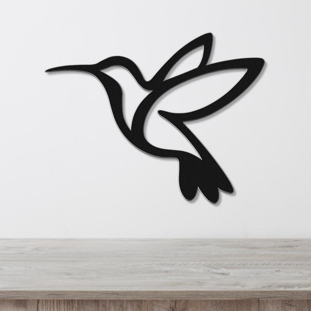 Black metal hummingbird wall art with a silhouette cutout design, mounted on a white wall above a light wood surface.