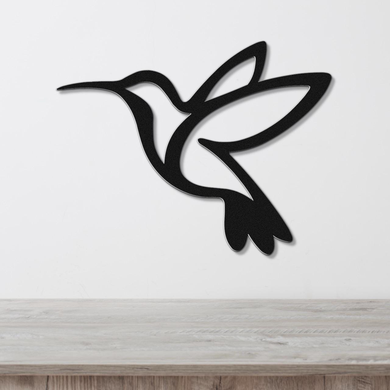Black metal hummingbird wall art with a silhouette cutout design, mounted on a white wall above a light wood surface.
