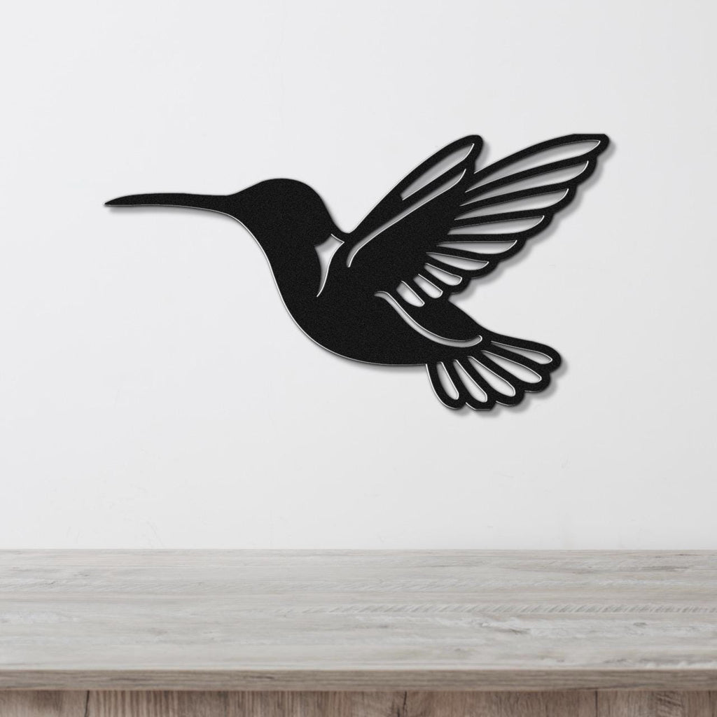 Black metal hummingbird wall art with wing and tail cutouts, mounted on a white wall above a light wood surface.