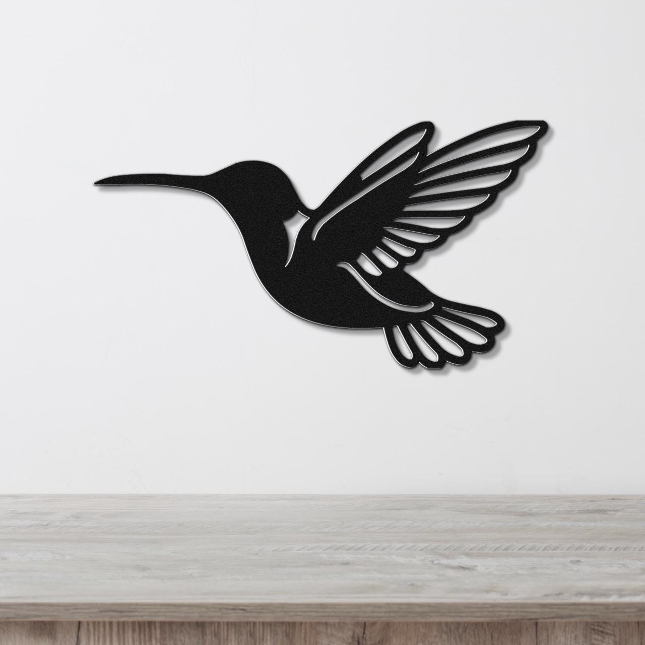 Black metal hummingbird wall art with wing and tail cutouts, mounted on a white wall above a light wood surface.