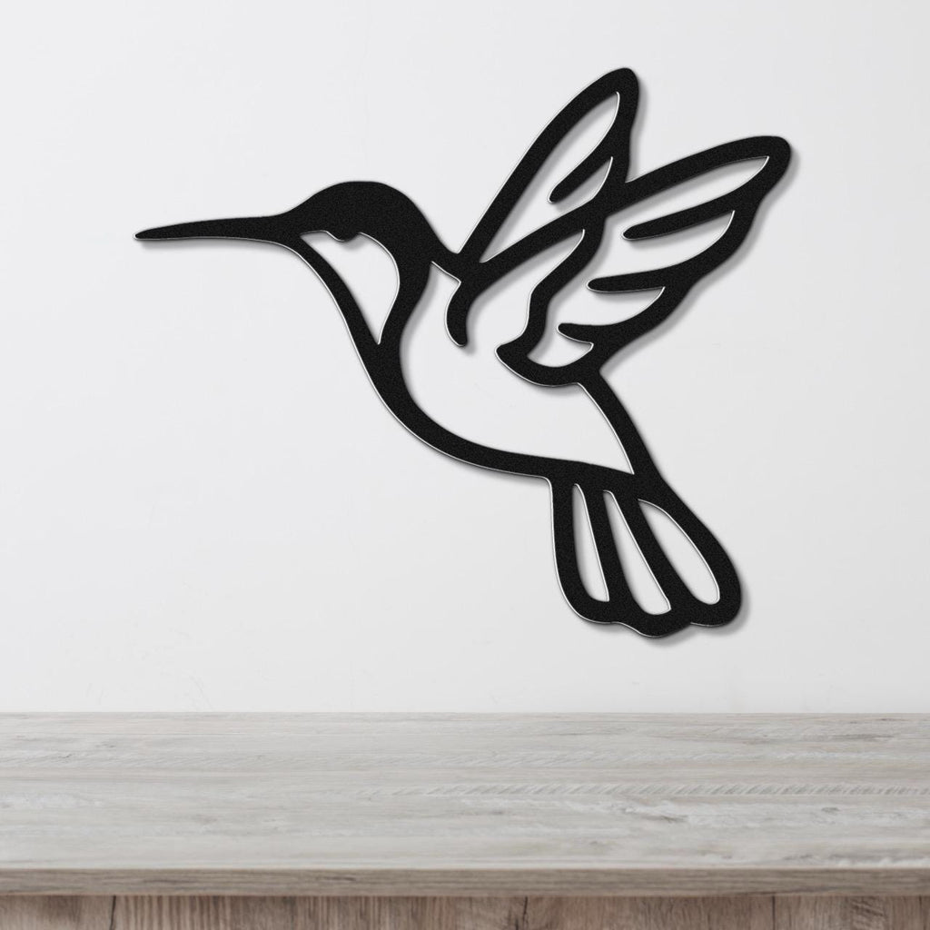 Black metal hummingbird wall art with wing and tail cutouts, mounted on a white wall above a light wood surface.