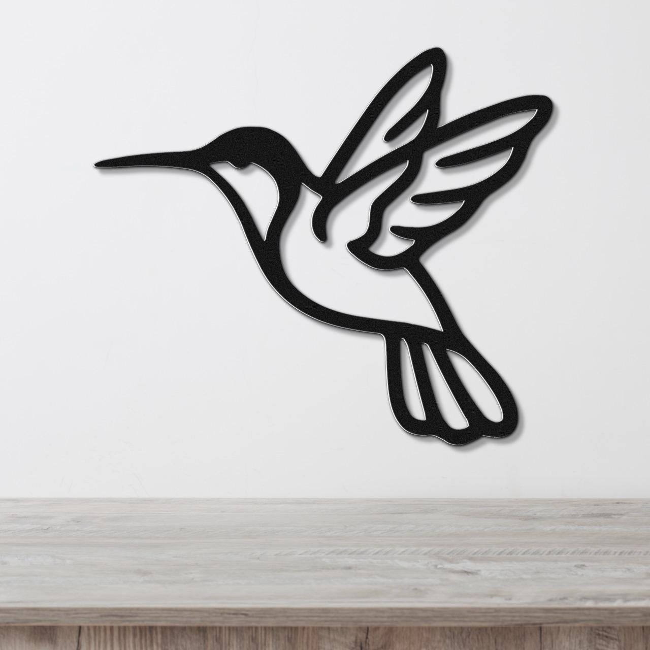 Black metal hummingbird wall art with wing and tail cutouts, mounted on a white wall above a light wood surface.
