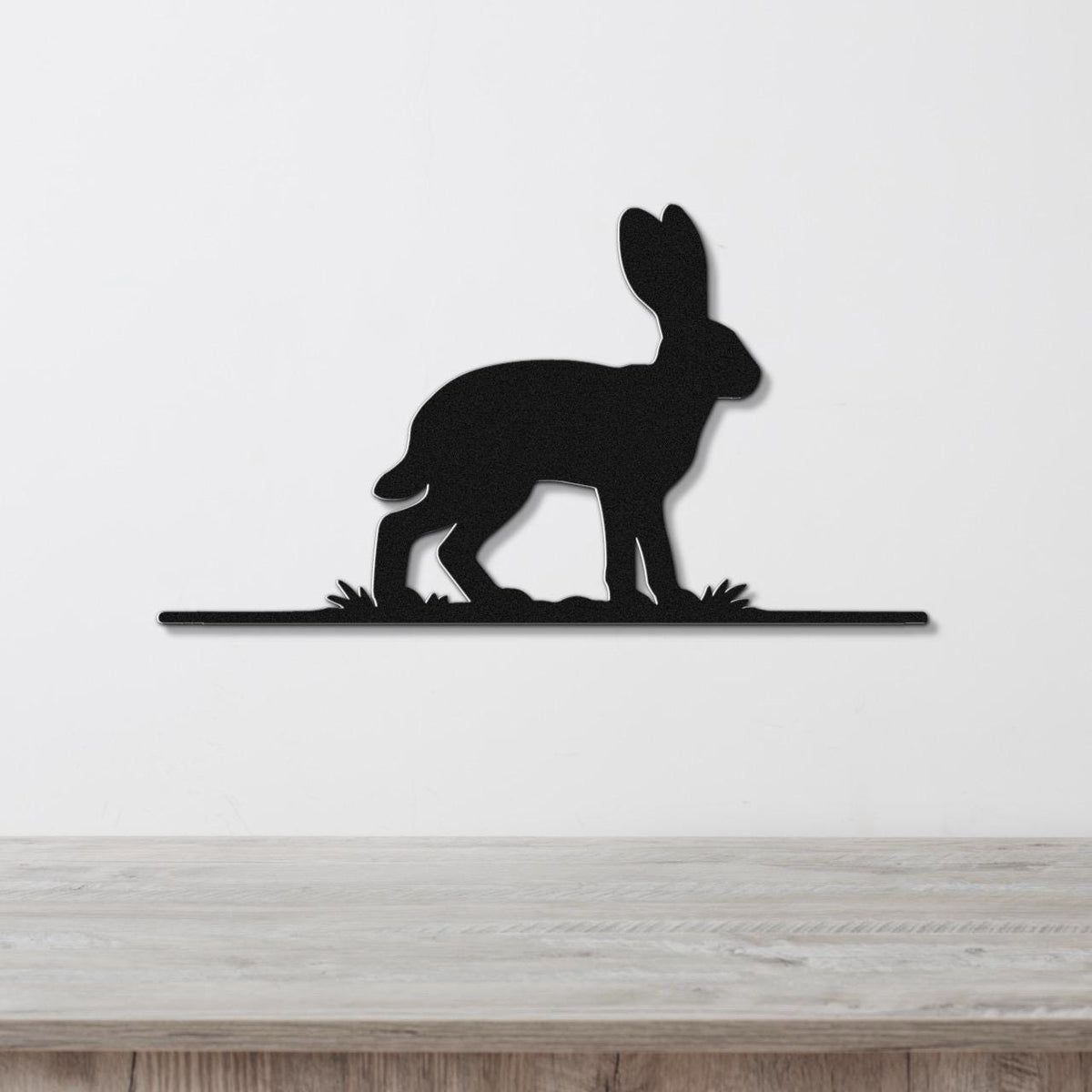 Black metal jackrabbit wall art silhouette with grass cutouts, mounted on a white wall above a wooden surface.
